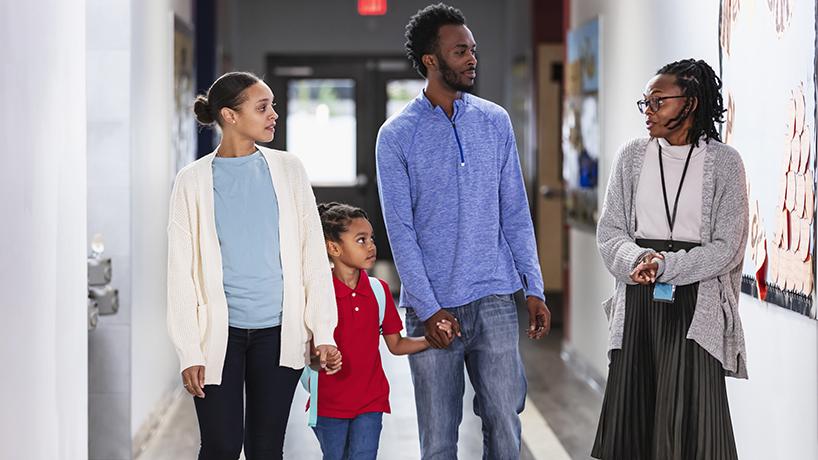 Parents bringing girl to school, with teacher in hallway